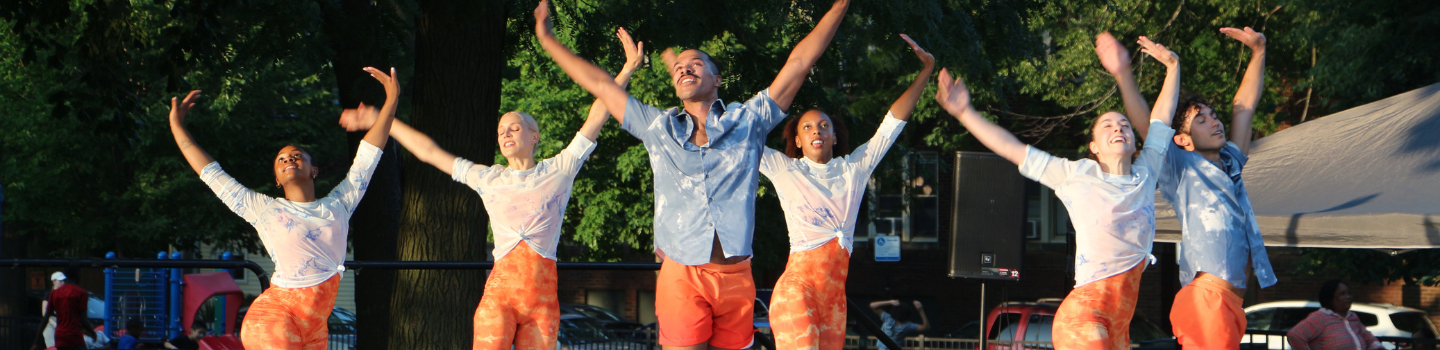 Six dancers perform outdoors with arms raised. They wear orange pants or shorts and sheer white or blue tops.
